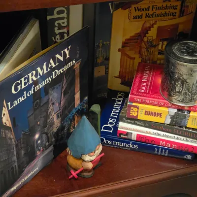 Sale Photo Thumbnail #220: Collection of hardcover and paperback books, an embossed metal trinket tin, and a carved wooden gnome figurine on a shelf.