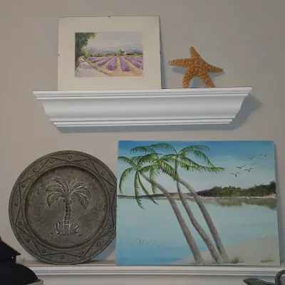 Sale Photo Thumbnail #174: Collection of tropical and coastal decor items, including a palm tree oil painting on canvas, a embossed metal plate with a palm motif, a framed lavender field print, and a dried starfish displayed on white floating wall shelves.
