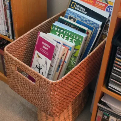 Sale Photo Thumbnail #119: Woven rattan storage basket filled with nature guidebooks, situated between wooden shelving units with DVDs and CDs.