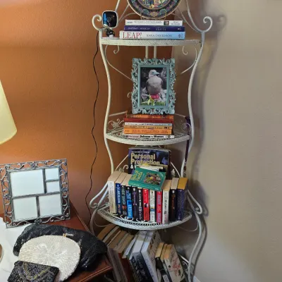 Sale Photo Thumbnail #141: White wrought iron four-tier corner baker's rack holding books, framed pictures, and decorative items alongside a side table displaying beaded purses.