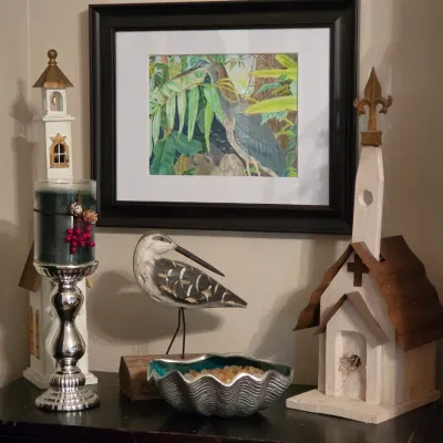 Sale Photo Thumbnail #192: Collection of coastal decor items, including a framed heron watercolor print, carved shorebird figurine, decorative birdhouses, and a clam shell bowl on a dark tabletop.