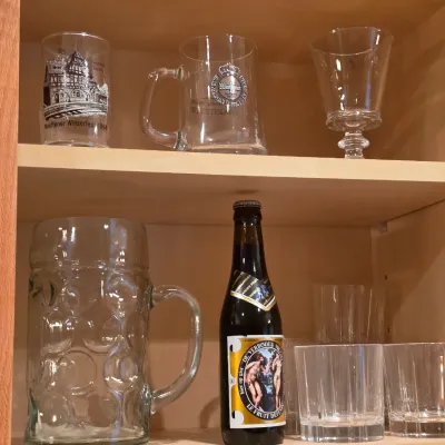 Sale Photo Thumbnail #20: Collection of glassware and a vintage bottle of De Verboden Vrucht beer displayed inside a wooden cabinet.