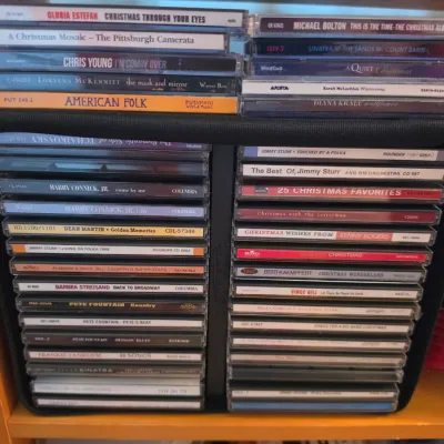 Sale Photo Thumbnail #115: Collection of assorted audio CDs in jewel cases, featuring holiday, folk, jazz, and pop music, stored in a black organizer rack.