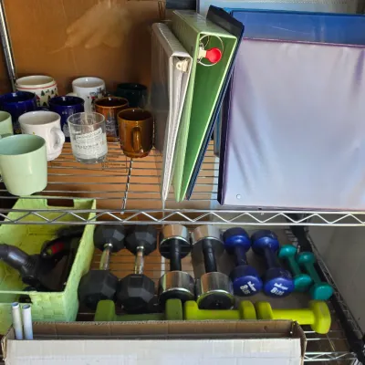 Sale Photo Thumbnail #307: Wire storage shelving holding an assortment of ceramic coffee mugs, three-ring binders, and various hand weights.