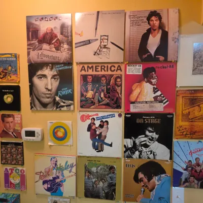 Sale Photo Thumbnail #211: Collection of vintage vinyl record albums and 45 RPM sleeves displayed on a wall, featuring artists like Bruce Springsteen, Elvis Presley, The Monkees, and Kansas.