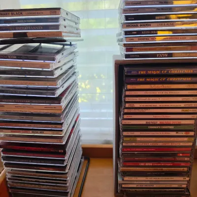 Sale Photo Thumbnail #114: Large collection of compact discs in jewel cases, featuring various pop, country, and holiday music artists stacked on a desk and in a storage rack.