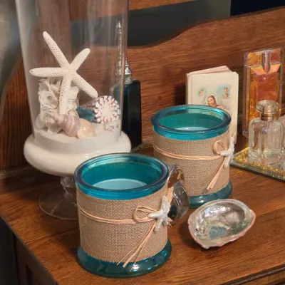 Sale Photo Thumbnail #187: Coastal-themed dresser decor, featuring turquoise glass candle holders wrapped in burlap, a glass terrarium filled with shells and starfish, an abalone shell, and assorted perfume bottles on a wooden vanity.
