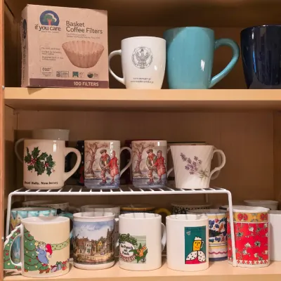 Sale Photo Thumbnail #22: Collection of assorted ceramic coffee mugs and a box of coffee filters stored on wooden cabinet shelves with a metal wire organizer.