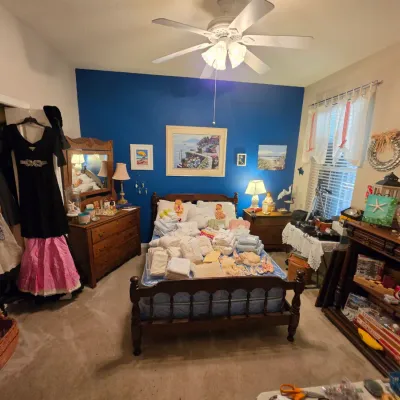 Sale Photo Thumbnail #166: Bedroom filled with estate sale items, including a wooden spindle bed piled with linens, a matching dresser with mirror, clothing hanging in the closet, framed coastal artwork, and board games.