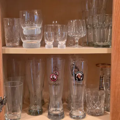 Sale Photo Thumbnail #18: Collection of glassware stored in a wooden cabinet, including branded German beer glasses, crystal brandy snifters, and cut glass tumblers.