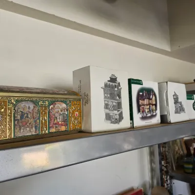 Sale Photo Thumbnail #288: Collection of Department 56 Heritage Village buildings in original boxes alongside a decorative ornate metal tin on a metal shelving unit.