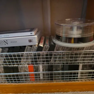 Sale Photo Thumbnail #118: Wire storage basket shelf filled with cassette tapes, boxed sets, and a spindle of blank recordable CDs.