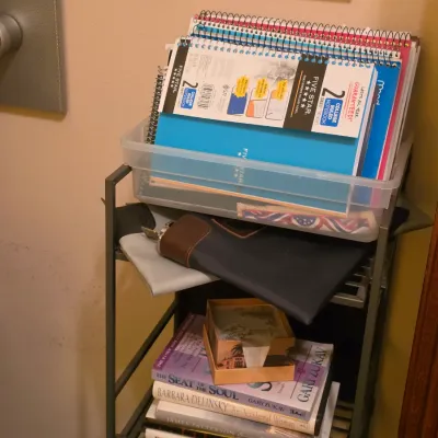 Sale Photo Thumbnail #225: A small multi-tiered utility cart holding plastic bins filled with spiral notebooks, padded document holders, and a stack of paperback books.