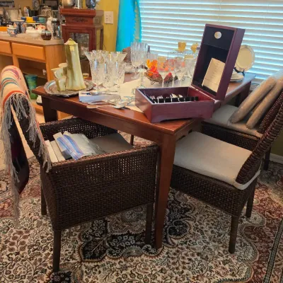 Sale Photo Thumbnail #104: Wooden dining table display set with woven dark wicker chairs, featuring crystal stemware, a boxed silverware set, and decorative tableware.