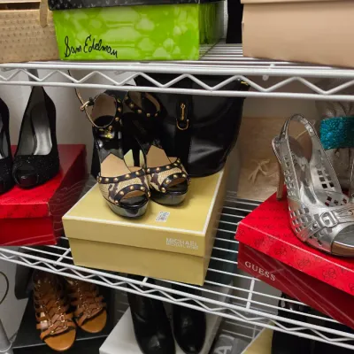 Sale Photo Thumbnail #149: Assortment of women's designer shoes including Guess black heels, Michael Kors studded snake-print sandals, and silver strappy heels displayed on shoe boxes on wire shelving.