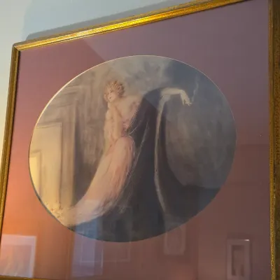 Sale Photo Thumbnail #139: Framed Art Deco oval print after Louis Icart depicting an elegant woman smoking a cigarette, set in a gold-painted frame with a wide mauve mat.