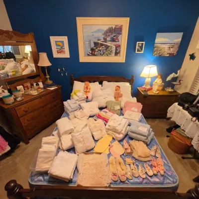Sale Photo Thumbnail #178: Bedroom filled with vintage linens, towels, baby clothes, and shoes displayed on a double bed, flanked by antique wooden dressers, table lamps, and framed coastal wall art.