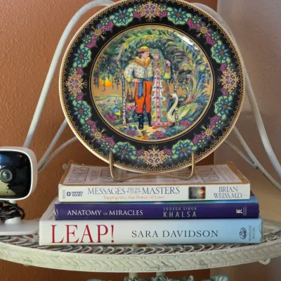 Sale Photo Thumbnail #142: Russian fairytale decorative collector plate displayed on a stand atop a stack of hardcover books, alongside a compact white security camera.