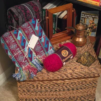 Sale Photo Thumbnail #190: Woven wicker trunk topped with colorful Southwestern-style fringe throws, a skein of Lion Brand Homespun yarn, a small copper pitcher, and a wooden mirror frame.