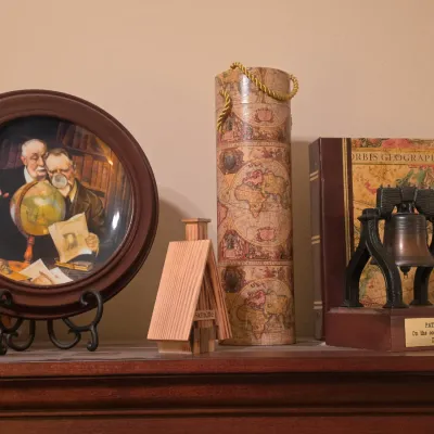 Sale Photo Thumbnail #214: Collection of decorative collectibles including a framed Norman Rockwell collector plate, a vintage map-pattern cylinder container, a small wooden cabin figurine, and a commemorative brass Liberty Bell desk plaque.