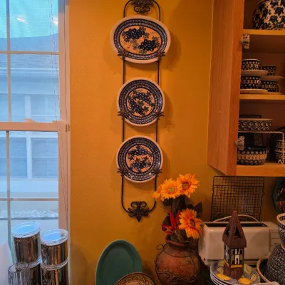 Sale Photo Thumbnail #41: A wall-mounted wrought iron plate rack displaying three Polish stoneware decorative plates with grape motifs, shown in a kitchen setting alongside various Polish pottery pieces.