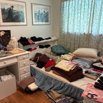 Sale Photo Thumbnail #3: Bedroom featuring a single bed covered with assorted folded linens and clothing, a white writing desk, framed artwork, and floral curtains.