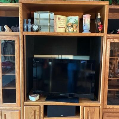 Sale Photo Thumbnail #9: Large light oak entertainment center featuring a Sony television, lower storage cabinets, glass-door display sections, and various decor items on the upper shelf.
