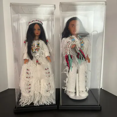 Pair of vintage Native American style fashion dolls dressed in white knit fringe outfits with beaded jewelry, displayed in clear acrylic...