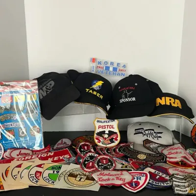 Collection of firearm, military, and locksmith memorabilia, including baseball caps, embroidered patches, decal stickers, and a packaged...