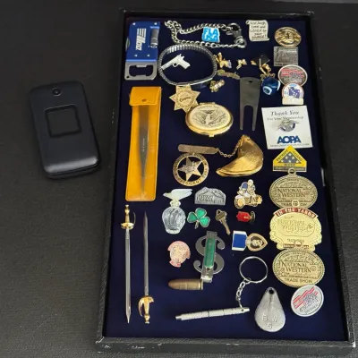 Collection of assorted vintage small collectibles and pins displayed in a tray next to a black flip phone, including lapel pins,...
