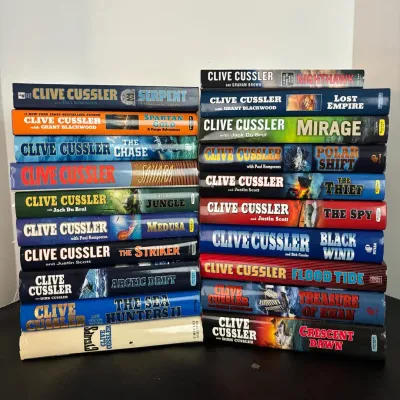 Collection of 20 Clive Cussler thriller and adventure hardcover novels, stacked in two piles.