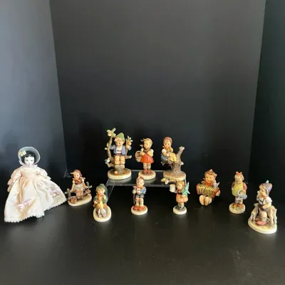 Collection of vintage Goebel Hummel porcelain figurines displayed alongside a porcelain doll in a pink dress.