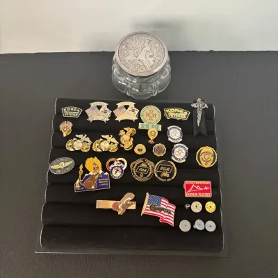 Collection of military, police, and organizational lapel pins displayed on a black velvet tray, accompanied by an embossed glass trinket...