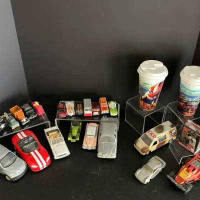 Collection of assorted die-cast toy vehicles and collectibles, featuring Hot Wheels and Matchbox cars along with two graphic souvenir cups.