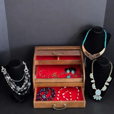 Vintage wooden roll-top jewelry box with red velvet-lined drawers, displayed alongside costume necklaces on black velvet busts.