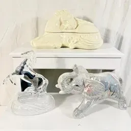 Sale Photo Thumbnail #50: Collection of three animal figurines, including a clear glass rearing horse, a clear glass trunk-up elephant, and a cream ceramic reclining elephant lidded box.