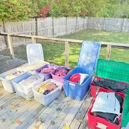 Sale Photo Thumbnail #84: Collection of plastic storage totes and bins filled with folded clothing and textiles, arranged on an outdoor wooden deck.