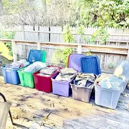 Sale Photo Thumbnail #83: Collection of large plastic storage totes filled with assorted clothing and household items, arranged outdoors on a wooden deck.