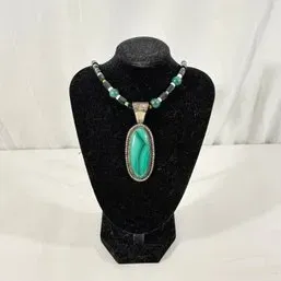 Sale Photo Thumbnail #251: Southwestern-style beaded necklace featuring a large oval green malachite cabochon pendant set in sterling silver on a black velvet display stand.