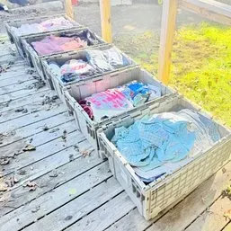 Sale Photo Thumbnail #79: Row of large plastic storage crates filled with assorted folded clothing and linens displayed outdoors on a wooden deck.