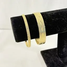 Sale Photo Thumbnail #250: Pair of gold-tone metal bangle bracelets featuring brushed finish textures and carved starburst details, displayed on a black velvet stand.