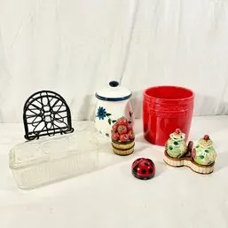 Sale Photo Thumbnail #69: Assorted kitchenware including a red utensil crock, floral ceramic canister, clear glass butter dish, black metal napkin holder, novelty salt and pepper shakers, and small decorative figurines.