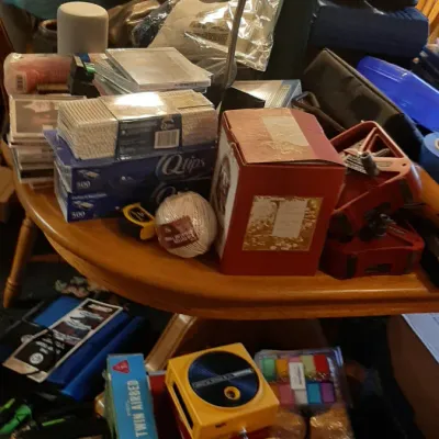 A wooden table densely packed with household items, including a metal clamp light, Q-tips boxes, a boxed portable record player, a red...