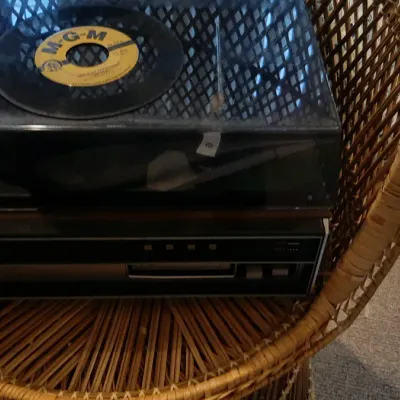 Vintage stereo turntable component system with tinted dust cover resting on a wicker chair, featuring a 45 RPM vinyl record on top.