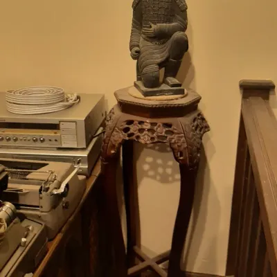 Replication terracotta warrior figurine displayed atop a tall carved Asian-style plant stand, alongside vintage electronics and a manual...