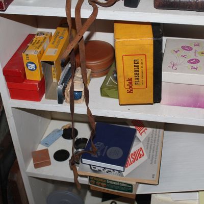 Sale Photo Thumbnail #88: Collection of vintage photography accessories and small boxes, including a Kodak Flasholder, General Electric flashbulbs, and leather camera straps on white shelving.