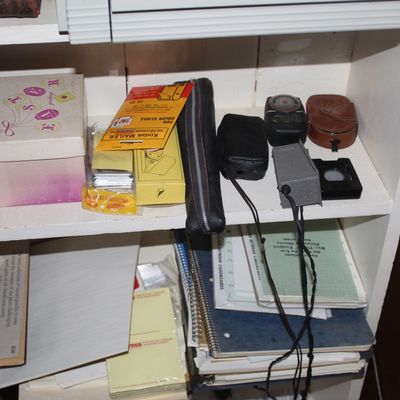 Sale Photo Thumbnail #89: A collection of vintage photography accessories and miscellany on white shelving, including Kodak mailers, flash accessories, leather cases, and small boxes.