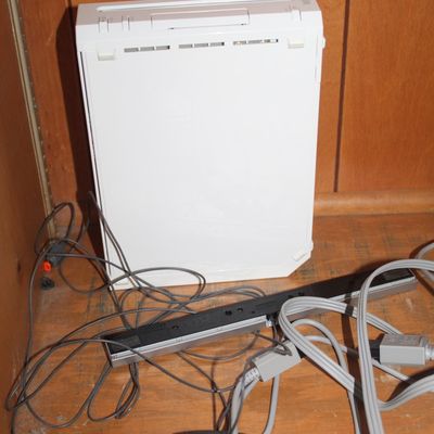 Sale Photo Thumbnail #70: White Nintendo Wii console with a sensor bar, power and AV cables, resting inside a wooden cabinet.