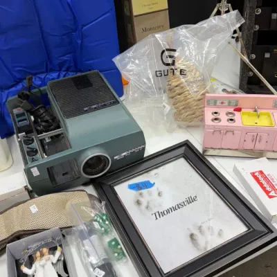 Assorted household items, including a vintage slide projector, Thomasville framed print, pink toy kitchen sink, and decorative accessories.