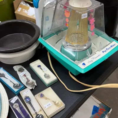 Vintage Suzy Homemaker electric Pop Corn Maker alongside boxed souvenir spoons and various household kitchenware.
